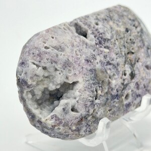 May include: A cylindrical lepidolite specimen featuring a rough, mottled purple and white exterior with a prominent, hollowed-out geode cavity filled with small, sparkling crystalline structures and natural surface pits throughout the stone.