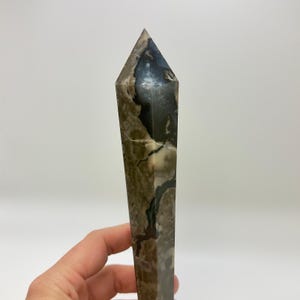 May include: A polished stone tower with a pointed top, featuring a marbled pattern of dark charcoal, earthy brown, and creamy white tones. The surface displays natural veining and organic color transitions throughout the elongated crystal form.