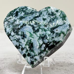 May include: A heart-shaped moss agate stone featuring a polished surface with intricate dark green, forest green, and black dendritic inclusions set against a translucent light gray and white background, displayed on a clear plastic stand.