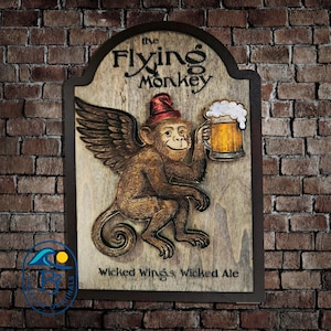 May include: Wooden sign featuring a winged monkey holding a beer mug. The sign reads "The Flying Monkey" and "Wicked Wings, Wicked Ale." The monkey wears a red hat and has a smiling expression. The sign is framed in dark brown and mounted on a brick wall.