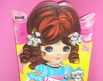 Könnte beinhalten: Vintage "Holly a Costume Paper Doll" Box. Die Box zeigt eine Cartoon-Illustration eines Mädchens in einem blauen Kleid, das ein Schild und ein Kätzchen hält. Die Box hat einen gelben Hintergrund mit rosa Akzenten und braunen Haaren.