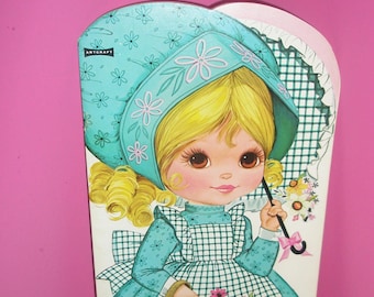Könnte beinhalten: Ein Vintage-Set mit Amy Jo Papierpuppen. Die Illustration zeigt ein Cartoon-Mädchen in einem türkisfarbenen Kleid und einer Haube, das einen Blumenkorb und einen Regenschirm hält. Das Design hat einen Retro-Stil mit rosa Hintergrund und dem Text "Amy Jo Paper Dolls".