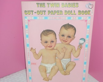 Könnte beinhalten: Ein Vintage-Papierpuppenbuch mit dem Titel "The Twin Babies Cut-Out Paper Doll Book". Das Cover zeigt Illustrationen von zwei Babys, eines mit einer Rassel. Das Buch hat einen rosa Hintergrund mit dekorativen Bordüren und Text.