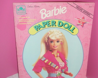 May include: A pink Barbie Paper Doll Deluxe Edition box from Golden Books. The box features a portrait of Barbie in a pink outfit with a blue belt and a colorful necklace. The box includes a paper doll, precut fashions, and accessories.