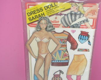 Könnte beinhalten: Ein Vintage-Papierpuppen-Set mit dem Titel "Dress Doll Sarah" in einer Plastikverpackung. Das Set enthält eine Papierpuppe mit verschiedenen Outfits, darunter einen Pullover, einen Rock und Accessoires. Die Verpackung zeigt Illustrationen der Puppe und der Outfits.