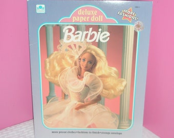 May include: A vintage Barbie Deluxe Paper Doll box. The box is light blue with the word "Barbie" in pink. The doll is wearing a white dress. The box reads "MORE FASHIONS!" and "more precut clothes fashions to finish storage envelope."