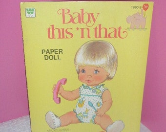 May include: A vintage "Baby this 'n that" paper doll set. The cover is yellow with a cartoon illustration of a baby doll holding a pink telephone. The doll is wearing a blue and white patterned outfit. The text "Baby this 'n that" is in pink.