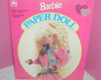 May include: A pink Barbie Paper Doll box featuring a blonde doll illustration in a pink dress and denim jacket. The box has the Barbie logo and the words "Paper Doll" in a large, circular design. Two glittery hearts are in the upper right corner.