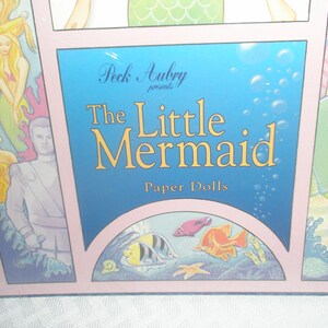 Op de afbeelding: Een doos voor "The Little Mermaid" papieren poppen. De doos heeft een blauwe achtergrond met bubbels en de titel in gouden letters. Illustraties van zeemeerminnen en zeedieren omringen de titel. De doos is roze omlijst.