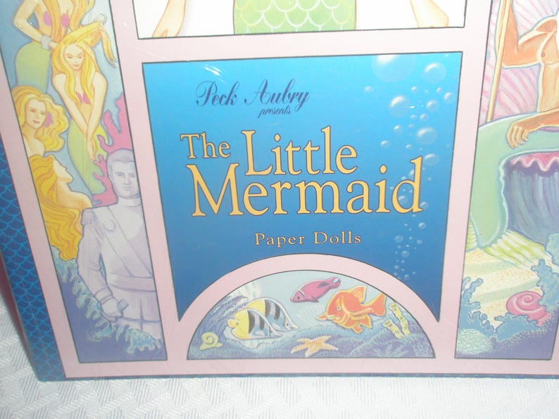 Op de afbeelding: Een doos voor "The Little Mermaid" papieren poppen. De doos heeft een blauwe achtergrond met bubbels en de titel in gouden letters. Illustraties van zeemeerminnen en zeedieren omringen de titel. De doos is roze omlijst.
