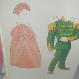 Puede incluir: Un conjunto de mu&ntilde;ecos de papel que incluye un vestido color melocot&oacute;n con un tocado a juego y un ramo de flores, junto a un uniforme militar verde con charreteras doradas, adornos trenzados y detalles de llamas amarillas en las mangas.