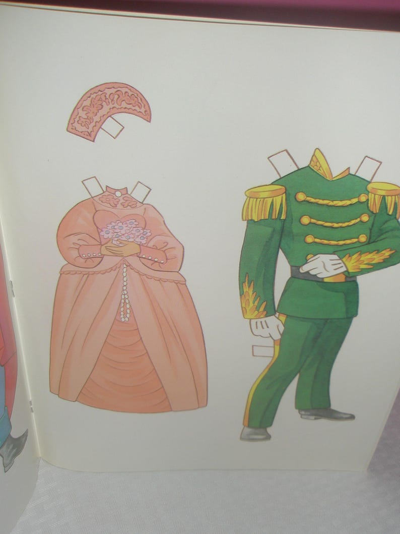 Puede incluir: Un conjunto de mu&ntilde;ecos de papel que incluye un vestido color melocot&oacute;n con un tocado a juego y un ramo de flores, junto a un uniforme militar verde con charreteras doradas, adornos trenzados y detalles de llamas amarillas en las mangas.