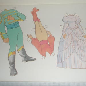 Puede incluir: Una hoja de mu&ntilde;eca de papel con tres elementos: una figura con uniforme militar color verde azulado con adornos dorados y botas grises, una blusa rosa de manga larga con cintura rosa y un vestido blanco largo hasta el suelo con capas de volantes.