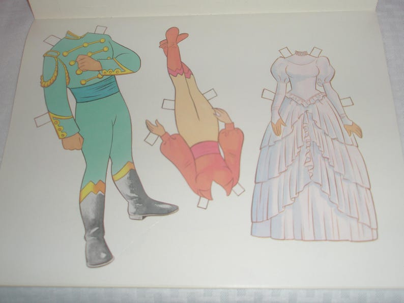 Puede incluir: Una hoja de mu&ntilde;eca de papel con tres elementos: una figura con uniforme militar color verde azulado con adornos dorados y botas grises, una blusa rosa de manga larga con cintura rosa y un vestido blanco largo hasta el suelo con capas de volantes.