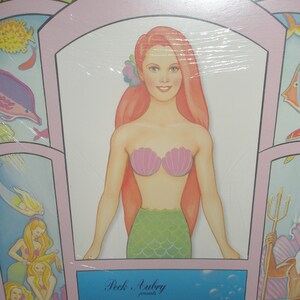 Op de afbeelding: Een verpakt product met een zeemeermin illustratie. De zeemeermin heeft lang rood haar, een roze schelpen top en een groene schubbenstaart. De tekst "The Little Mermaid" wordt weergegeven op een blauwe achtergrond.