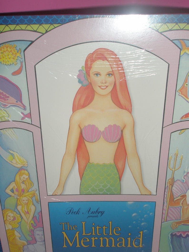 Op de afbeelding: Een verpakt product met een zeemeermin illustratie. De zeemeermin heeft lang rood haar, een roze schelpen top en een groene schubbenstaart. De tekst "The Little Mermaid" wordt weergegeven op een blauwe achtergrond.