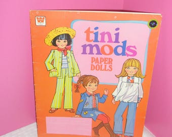 Puede incluir: Libro de muñecas de papel vintage "tini mods". La portada presenta ilustraciones de tres chicas de dibujos animados con atuendos de estilo mod. El libro es naranja con un diseño rosa y azul y las palabras "tini mods" en rosa y morado.