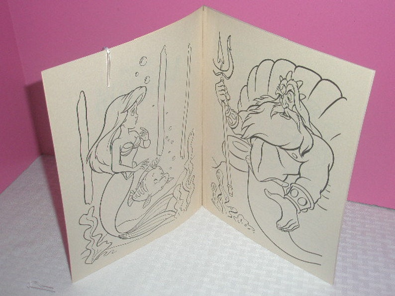 May include: A coloring book open to two pages, featuring line art illustrations. One page shows a mermaid with a fish, the other shows a bearded man on a throne. The images are in black and white.