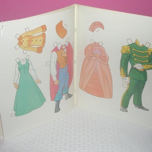 Puede incluir: Un libro de mu&ntilde;ecas de papel con dos figuras ilustradas y varios atuendos. Una figura viste un uniforme militar verde con charreteras doradas, mientras que la otra lleva un chaleco y una capa. Se incluye un vestido de color melocot&oacute;n.