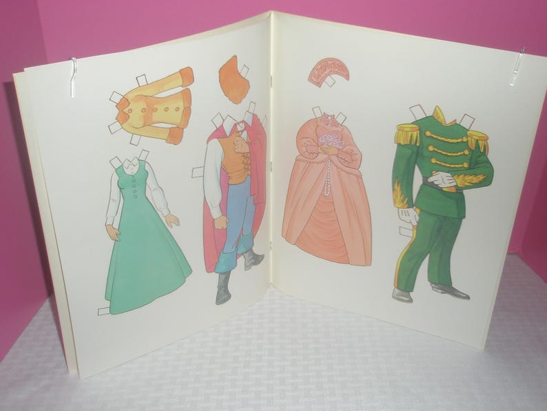 Puede incluir: Un libro de mu&ntilde;ecas de papel con dos figuras ilustradas y varios atuendos. Una figura viste un uniforme militar verde con charreteras doradas, mientras que la otra lleva un chaleco y una capa. Se incluye un vestido de color melocot&oacute;n.