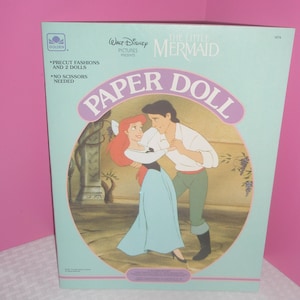 Puede incluir: Un libro de mu&ntilde;ecas de papel azul claro de Walt Disney La Sirenita con una ilustraci&oacute;n de Ariel y el Pr&iacute;ncipe Eric bailando. El texto de la portada incluye Precut Fashions and 2 Dolls, No Scissors Needed y el logo de Golden arriba.