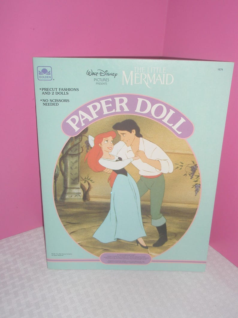 Puede incluir: Un libro de mu&ntilde;ecas de papel azul claro de Walt Disney La Sirenita con una ilustraci&oacute;n de Ariel y el Pr&iacute;ncipe Eric bailando. El texto de la portada incluye Precut Fashions and 2 Dolls, No Scissors Needed y el logo de Golden arriba.