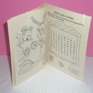 May include: Open coloring book featuring line art of Ariel and Flounder from The Little Mermaid. The book includes a word search puzzle titled "Grotto See-A-Word" and is held open with silver paper clips.