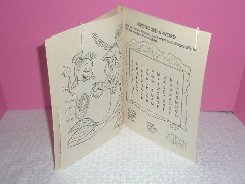 May include: Open coloring book featuring line art of Ariel and Flounder from The Little Mermaid. The book includes a word search puzzle titled "Grotto See-A-Word" and is held open with silver paper clips.