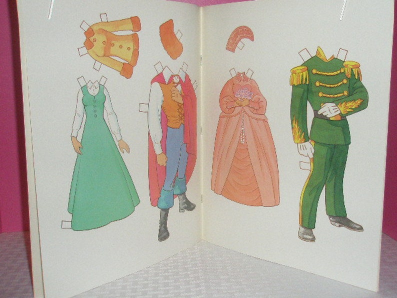 May include: An open book featuring paper dolls with various outfits. The outfits include a green dress, a yellow coat, a prince's attire with a red cape, a pink gown, and a green military uniform. The paper dolls are designed with tabs for easy removal.