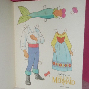 May include: A vintage paper doll set featuring characters from Disney's The Little Mermaid. The set includes a mermaid tail, a dress, and a male character with blue pants and a white shirt. The title "The Little Mermaid" is printed in gold.