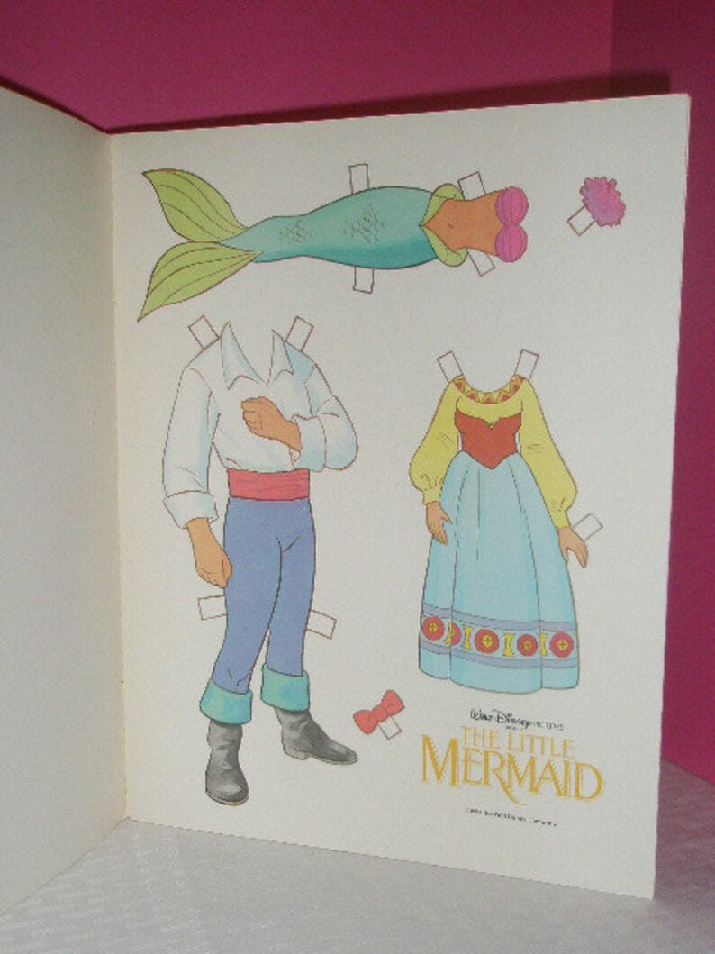 May include: A vintage paper doll set featuring characters from Disney's The Little Mermaid. The set includes a mermaid tail, a dress, and a male character with blue pants and a white shirt. The title "The Little Mermaid" is printed in gold.