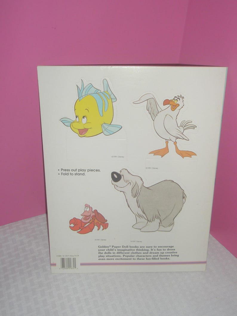 Puede incluir: Una contraportada rectangular blanca de un libro de mu&ntilde;ecas de papel con ilustraciones coloridas de un pez amarillo, una gaviota blanca, un cangrejo rojo y un perro gris. El texto incluye Press out play pieces, Fold to stand y una descripci&oacute;n.