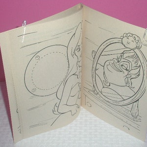 May include: An open coloring book featuring black and white illustrations. One page shows a fairy-like figure, and the facing page depicts a character in a circular frame. The pages are held open with clear clips.