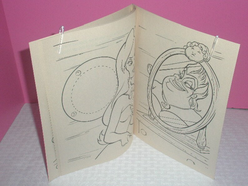 May include: An open coloring book featuring black and white illustrations. One page shows a fairy-like figure, and the facing page depicts a character in a circular frame. The pages are held open with clear clips.