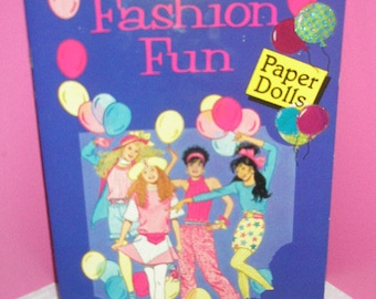 Puede incluir: Un libro azul titulado "Fashion Fun" con ilustraciones de muñecas de papel con trajes coloridos. La portada presenta el texto "Paper Dolls" en un cuadrado amarillo y está decorada con globos.