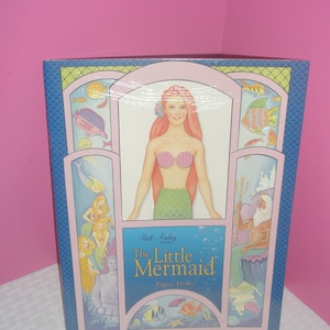 Op de afbeelding: Een doos met "The Little Mermaid Paper Dolls" met illustraties van zeemeerminnen en zeedieren. De doos heeft een blauwe achtergrond met roze en groene accenten en de titel staat in een gestileerd lettertype.