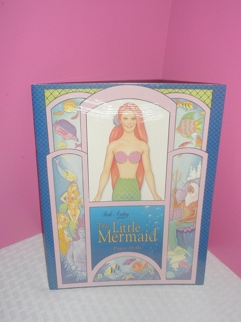 Op de afbeelding: Een doos met "The Little Mermaid Paper Dolls" met illustraties van zeemeerminnen en zeedieren. De doos heeft een blauwe achtergrond met roze en groene accenten en de titel staat in een gestileerd lettertype.