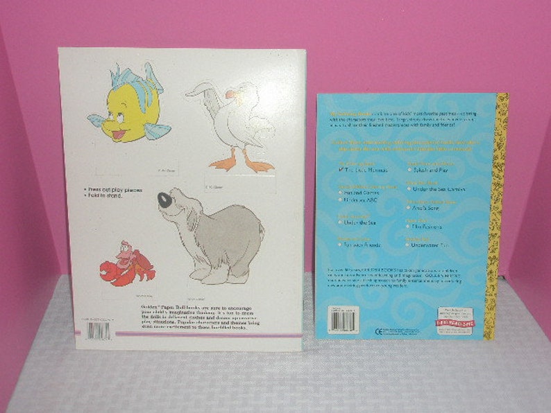 May include: Two children's books are displayed. One features illustrations of cartoon characters, including a yellow fish, a white seagull, a gray dog, and a red crab. The other book has a blue cover with gold trim and text.