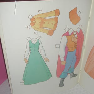 Puede incluir: Una p&aacute;gina de libro de mu&ntilde;ecas de papel que muestra una chaqueta amarilla con botones, un vestido verde con una camisa blanca de cuello, y una figura con chaleco amarillo, pantalones azules, botas negras y una capa roja larga, con una pieza de pelo naranja.