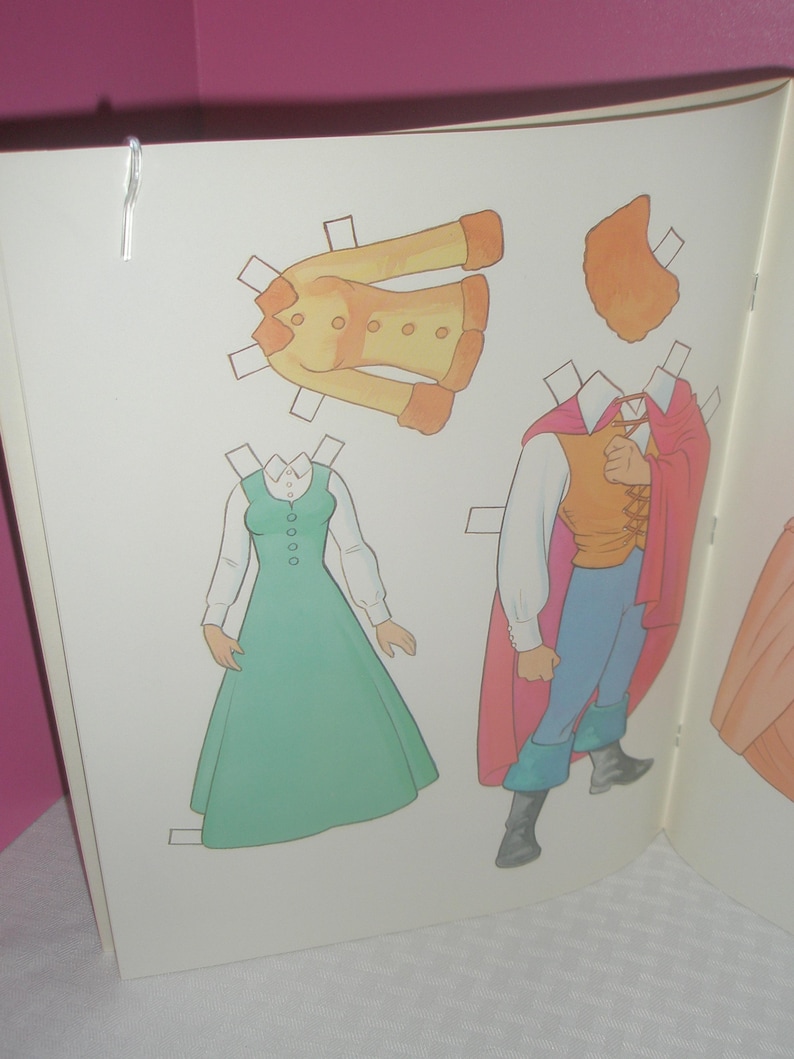 Puede incluir: Una p&aacute;gina de libro de mu&ntilde;ecas de papel que muestra una chaqueta amarilla con botones, un vestido verde con una camisa blanca de cuello, y una figura con chaleco amarillo, pantalones azules, botas negras y una capa roja larga, con una pieza de pelo naranja.