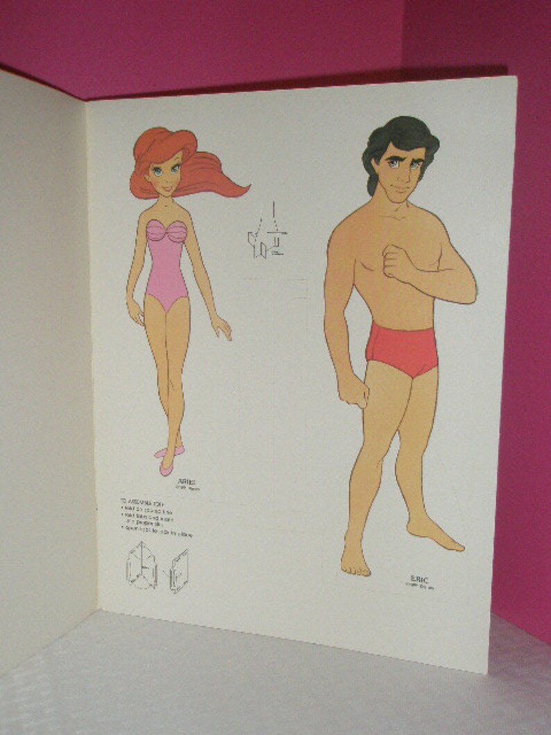 May include: A page from a vintage paper doll book featuring illustrations of Ariel and Eric. Ariel is in a pink swimsuit, and Eric is in red swim trunks. The characters are drawn in a cartoon style.