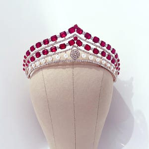 Op de afbeelding: Een gouden tiara met drie rijen rode en heldere strass steentjes en een rij witte parels.