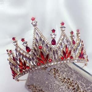 Pode incluir: Uma coroa dourada com strass vermelhos e transparentes. A coroa é feita de metal e tem um topo pontudo.