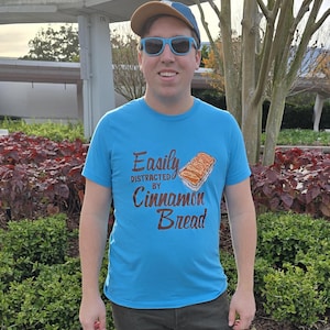 May include: A man wearing a blue t-shirt with the text "Easily Distracted By Cinnamon Bread" and a graphic of a loaf of cinnamon bread. He is wearing sunglasses and a brown baseball cap.