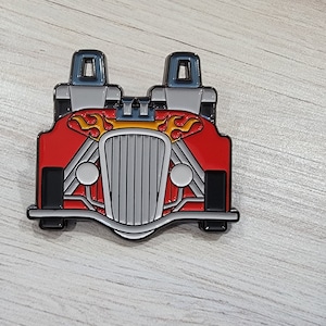 Puede incluir: Un pin de esmalte rojo y plateado de un coche de carreras vintage con llamas en el capó. El coche tiene dos asientos y una parrilla negra.