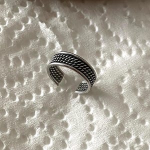Puede incluir: Un anillo de dedo del pie ajustable plateado con un diseño de cuerda trenzada. El anillo es abierto en la parte inferior y presenta un patrón detallado alrededor de la banda. El anillo se muestra sobre una superficie blanca texturizada.