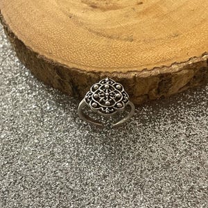 May include: A silver adjustable ring with an intricate, symmetrical design. The ring's central element showcases a detailed pattern of curves and dots. The ring is set against a sparkly, silver background and a wooden slice.