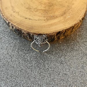 May include: A silver-colored ring featuring a detailed jaguar head design. The ring has an adjustable band and is set against a textured gray surface and a wooden slice.