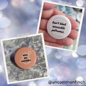 Könnte beinhalten: Zwei runde Anstecknadeln mit weißem Hintergrund und schwarzem Text. Die erste Nadel sagt "Make Shit Accessible" mit einem stilisierten Rollstuhl-Symbol. Die zweite Nadel sagt "Blockiere keine barrierefreien Wege."