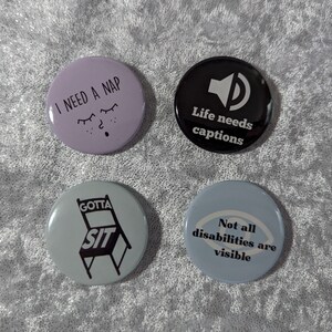 May include: Four round button pins with different messages. The first pin is purple with the text "I need a nap" and a cartoon eye with eyelashes. The second pin is black with a white speaker icon and the text "Life needs captions". The third pin is light blue with a black chair and the text "Gotta sit". The fourth pin is light blue with a white eye icon and the text "Not all disabilities are visible".